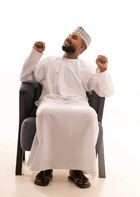 ايماءات السعادة والنشاط، مفهوم تمطيط الجسم والاسترخاء، بورتريه لرجل عربي عماني يرتدي الدشداشة والكمه يجلس على كرسي يقوم بحركات تمدد عضلات الجسم، الشعور بالراحة والنشاط، خلفية بيضاء