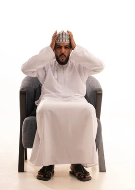 تعابير وجه تدل على حدوث مصيبة ما، بورتريه لرجل عربي عماني يرتدي الدشداشة والكمه يجلس على كرسي  يضع يده على رأسه تظهر عليه علامات الحيرة والوقوع في مشكلة، عدم القدرة على مسك زمام الأمور وفقدان السيطرة، خلفية بيضاء