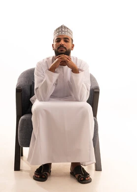 Contemplation, focus, and planning for the future, finding a solution to a problem, a portrait of an Arab Gulf Omani man wearing a dishdasha and kumma sitting on a chair with his hand under his chin in gestures of contemplation and thought, the concept of daydreaming, white background.