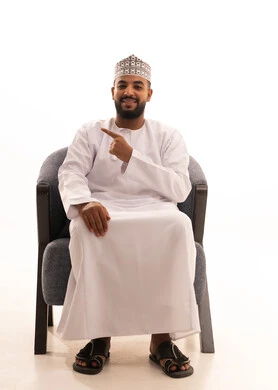 النظر الكاميرا بإيماءات السرور والبشاشة، بورتريه لرجل عربي خليجي عماني يرتدي الدشداشة و الكمة  يجلس على كرسي يشير بأصابعه الى هدف محدد، إشارات وحركات يدين تدل على هذا المكان (هنا)، تحديد موقع معين، خلفية بيضاء