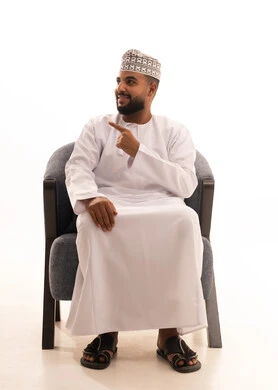 تحديد موقع معين، النظر الكاميرا شيء ما، بورتريه لرجل عربي خليجي عماني يرتدي الدشداشة و الكمة يجلس على كرسي يشير بأصابعه الى هدف محدد، إشارات وحركات يدين تدل على هذا المكان (هنا)، خلفية بيضاء