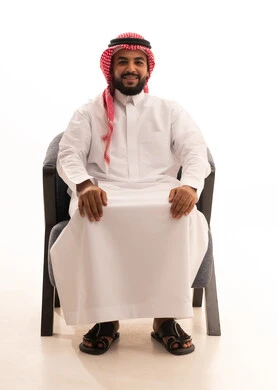 ايماءات الثقة بالنفس، الاهتمام بالأناقة والمظهر الخارجي، مفهوم القوة والرجولة والشياكة،بورتريه لرجل عربي خليجي سعودي يرتدي الثوب التقليدي والشماغ يجلس على كرسي ينظر الى الكاميرا بتعابير الفرح والسرور، خلفية بيضاء