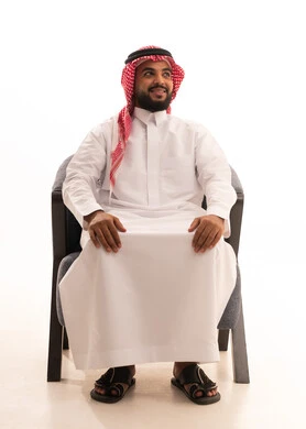 The concept of strength, masculinity, and elegance, the interest in style and appearance, a portrait of a Saudi Gulf Arab man wearing traditional attire and a shemagh sitting on a chair looking at something with expressions of joy and happiness, gestures of self-confidence, white background.