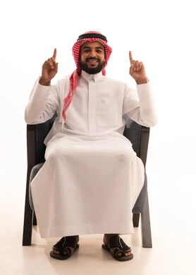 ايماءات السعادة و السرور ، اشارة رقم واحد،  بورتريه لرجل عربي خليجي سعودي يرتدي الثوب التقليدي والشماغ يجلس على كرسي يشير بإصبعه السبابة للاعلى إلى شيء محدد بتعابير هذا المكان هنا، خلفية بيضاء .