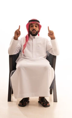 Expressions of astonishment and wonder, sign number one, a portrait of a Saudi Arabian Gulf man wearing traditional attire and a ghutrah sitting on a chair pointing with his index finger upwards to something specific with the expressions of this place here, white background.