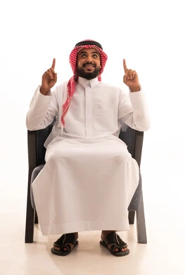 اشارة رقم واحد، النظر إلى الاعلى بإيماءات الفرح والسرور، بورتريه لرجل عربي خليجي سعودي يرتدي الثوب التقليدي والشماغ يجلس على كرسي يشير بإصبعه السبابة للاعلى إلى شيء محدد بتعابير هذا المكان هنا، خلفية بيضاء .