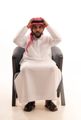تذكر شيء ما، الشعور بالدهشة والذهول, بورتريه لرجل عربي خليجي سعودي يرتدي الثوب التقليدي والشماغ يجلس على كرسي يضع يديه على رأسه بإيماءات التعجب والانبهار, النظر إلى الكاميرا بايماءات الصدمة, التفاجؤ من موقف محدد، خلفية بيضاء
