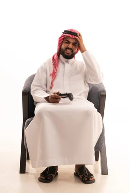 The hand is placed on the head with expressions of defeat, interacting and merging with video games, having a fun and entertaining time, using modern recreational electronic devices and technologies, a portrait of a Saudi Arabian Gulf man wearing traditional attire and a ghutrah sitting on a chair holding a game controller, with a white background.