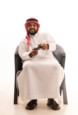 Enjoying modern technologies and technology, gestures of interaction and integration, a portrait of an Arab Gulf Saudi man wearing traditional thobe and ghutrah holding a game controller looking with expressions of joy, the concept of fun and entertainment, spending enjoyable time playing video games, white background.