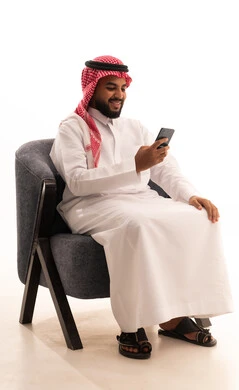 Communicating with individuals via mobile phone, browsing the internet and online shopping, using modern technologies and devices, chatting and conversations and meeting new people, and a portrait of a Saudi Arabian Gulf man wearing traditional attire and a ghutrah sitting on a chair using a mobile phone, with a white background.