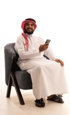 Looking at something with gestures of happiness and joy, browsing the internet and online shopping, using modern technologies and devices, communicating with individuals via mobile phone, chatting and conversations and getting to know each other, and a portrait of a Saudi Arabian Gulf man wearing traditional attire and a ghutrah sitting on a chair using a mobile phone, with a white background.