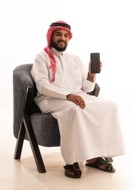 Display of mobile applications and services, mobile display with a blank black screen, use of advanced technology for digital electronic marketing, a portrait of a Saudi Arabian Gulf man wearing traditional attire and a ghutrah sitting on a chair looking at the camera with gestures of happiness and joy, white background.