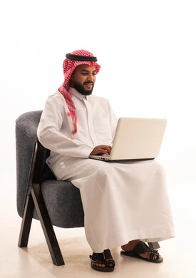 Managing and organizing business affairs through technical devices, gestures of integration and focus, a portrait of a Saudi Arabian Gulf man wearing traditional attire and a headscarf sitting on a chair working on a laptop, with a white background.