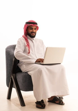 Gestures of happiness and joy, remote work, using advanced modern technology in professional life, a portrait of a Saudi Arabian Gulf man wearing traditional attire and a shemagh sitting on a chair working on a laptop, white background.