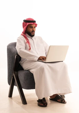 Remote work, using advanced modern technology in professional life, gestures of engagement and focus, a portrait of an Arab Gulf Saudi man wearing traditional attire and a headscarf sitting on a chair working on a laptop, white background.