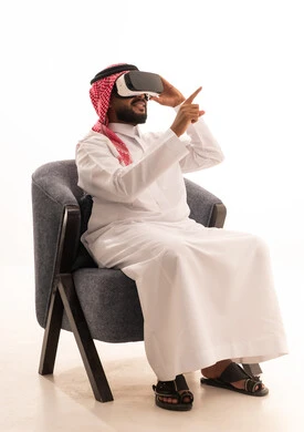 Pointing with the index finger at something, using modern technological devices, interacting and merging with virtual reality, seeing imaginary scenes, a portrait of a Saudi Arabian Gulf man wearing traditional attire sitting on a chair wearing VR glasses, spending leisure time in an enjoyable way, white background.