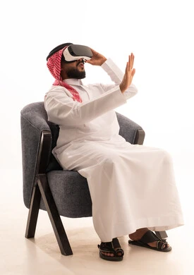Gestures of astonishment and surprise, the use of modern technological devices, interaction and immersion with virtual reality, seeing imaginary scenes, a portrait of an Arab Gulf Saudi man wearing traditional attire sitting on a chair wearing VR glasses, spending leisure time in an enjoyable way, white background.