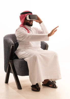 The use of modern technological devices, interaction and immersion with virtual reality, seeing imaginary scenes, a portrait of an Arab Gulf Saudi man wearing traditional attire sitting on a chair wearing VR glasses, spending leisure time in an enjoyable way, white background.