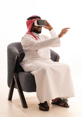 Seeing imaginary scenes, pointing with the index finger at something, using modern technological devices, interacting and merging with virtual reality, a portrait of a Saudi Arabian Gulf man wearing traditional attire sitting on a chair wearing VR glasses, spending leisure time enjoyably, white background.