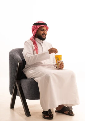 The concept of entertainment and leisure, having a healthy and light snack, spending free time watching entertaining movies and series, a portrait of a Gulf Arab Saudi man wearing traditional attire sitting on a chair and eating popcorn, white background.