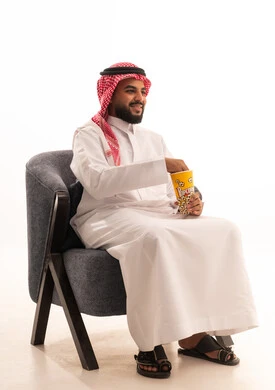 Eating a healthy and light snack, spending free time watching entertaining movies and series, a portrait of a Saudi Arabian Gulf man wearing traditional attire sitting on a chair and eating popcorn, white background.