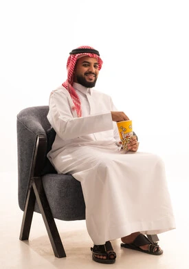 Spending free time watching entertaining movies and series, having a healthy and light snack, a portrait of a Gulf Arab Saudi man wearing traditional attire sitting on a chair and eating popcorn, white background.