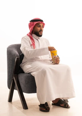 تعابير الدهشة والمفاجأة، مفهوم التسليه والترفيه، قضاء أوقات ممتعة في عطلة نهاية الاسبوع، أنشطة وفعاليات مسلية،  بورتريه لرجل عربي خليجي سعودي يرتدي الثوب التقليدي يجلس على كرسي ويأكل الفوشار، تناول الوجبات الخفيفة والسناك، خلفية بيضاء
