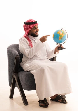 Identifying the location of Arab countries on the world map, research and exploration about the universe, a portrait of a Saudi Gulf Arab man wearing traditional attire sitting on a chair holding a globe model in his hand, the concept of trips and tourist tours, white background. Identifying the location of Arab countries on the world map, research and exploration about the universe, a portrait of a Saudi Gulf Arab man wearing traditional attire sitting on a chair holding a globe model in his hand, the concept of trips and tourist tours, white background.