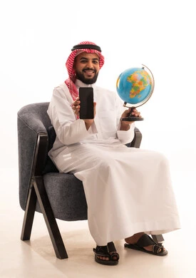 A blank black screen displayed on a mobile phone, the concept of the love for research and exploring continents in geography, a portrait of an Arab Gulf Saudi man wearing traditional attire sitting on a chair holding a globe in his hand, expressions of joy and happiness belonging to Arab countries, white background.