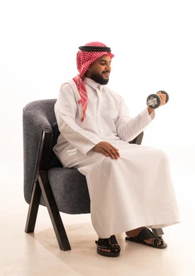 المحافظة على الصحة واللياقة الجسدية، ممارسة التمارين الرياضية مع الدمبل، تمارين رفع الأثقال (دمبلز)، بورتريه لرجل عربي خليجي سعودي يرتدي الثوب التقليدي يجلس على كرسي يقوم برفع الأوزان الحديدية الثقيلة، خلفية بيضاء