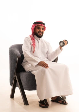 ممارسة التمارين الرياضية مع الدمبل، تمارين رفع الأثقال (دمبلز)، بورتريه لرجل عربي خليجي سعودي يرتدي الثوب التقليدي يجلس على كرسي يقوم برفع الأوزان الحديدية الثقيلة، المحافظة على الصحة واللياقة الجسدية، خلفية بيضاء