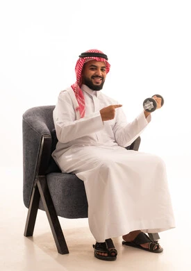 تمارين رفع الأثقال (دمبلز)،ممارسة التمارين الرياضية مع الدمبل، بورتريه لرجل عربي خليجي سعودي يرتدي الثوب التقليدي يجلس على كرسي يقوم برفع الأوزان الحديدية الثقيلة ويشير اليه، المحافظة على الصحة واللياقة الجسدية، خلفية بيضاء