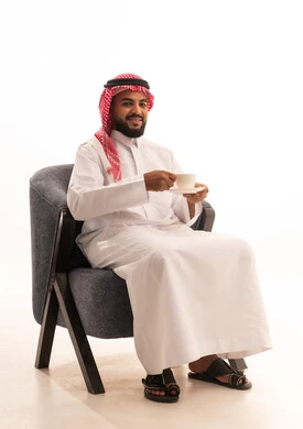 Sitting and enjoying hot drinks and warm winter herbs, expressions of happiness and joy, spending leisure time sipping the favorite drink, a portrait of an Arab Gulf Saudi man wearing traditional attire sitting on a chair holding a cup of coffee, white background.
