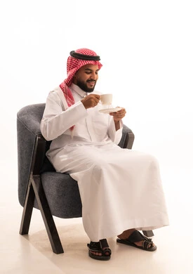 Expressions of happiness and joy, sipping the favorite drink, sitting and enjoying hot beverages and warm winter herbs, spending leisure time sipping the favorite drink, a portrait of a Saudi Arabian Gulf man wearing traditional attire sitting on a chair holding a cup of coffee, white background.