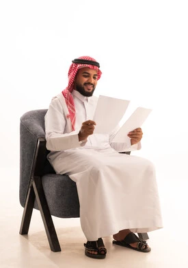 اعداد التقارير الخاصة بالعمل، مراجعة ومتابعة الخطط والمشاريع المكتبية بإيماءات التركيز، بورتريه لرجل عربي خليجي سعودي يرتدي الثوب التقليدي يجلس على كرسي يحمل بيده مجموعة من الاوراق البيضاء، التدقيق في قراءة الملاحظات، خلفية بيضاء