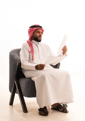 Reviewing and following up on office plans and projects with focused gestures, preparing work-related reports, a portrait of an Arab Gulf Saudi man wearing traditional attire sitting on a chair holding a stack of white papers, carefully reading notes, white background.