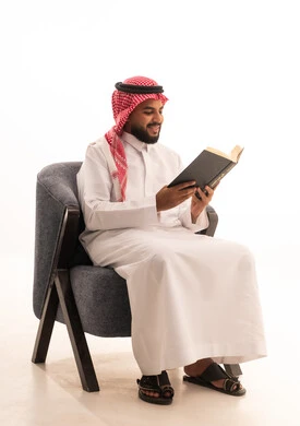 مفهوم التعلم والتفوق والاجتهاد، قضاء أوقات الفراغ بالأنشطة المفيدة، الاستمتاع بممارسة هواية المطالعة, بورتريه لرجل عربي خليجي سعودي يرتدي الثوب التقليدي والشماغ يجلس على كرسي  يقوم بقراءة الكتاب، خلفية بيضاء