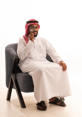 الفحص والاستكشاف والرؤية عن قرب، مفهوم توضيح الصورة، ايماءات تدل على السعادة والسرور، بورتريه لرجل عربي خليجي سعودي يرتدي الثوب التقليدي والشماغ يجلس على كرسي وينظر من خلال العدسة المكبرة، التدقيق والتركيز باستخدام الأدوات البصرية، خلفية بيضاء