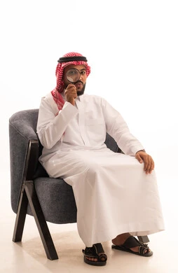 مفهوم توضيح الصورة، الفحص والاستكشاف والرؤية عن قرب، ايماءات تدل على السعادة والسرور، بورتريه لرجل عربي خليجي سعودي يرتدي الثوب التقليدي والشماغ يجلس على كرسي وينظر من خلال العدسة المكبرة، التدقيق والتركيز باستخدام الأدوات البصرية، خلفية بيضاء