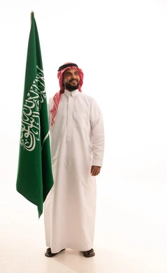 الاحتفال بذكرى اليوم الوطني السعودي 8 ربيع الأول, يوم العلم 11مارس،الفخر و الاعتزاز بالانتماء للمملكة, بورتريه لرجل عربي سعودي خليجي مبتسم يرتدي الشماغ والثوب التقليدي يحمل بيده علم السعودية , الجسم بالطول الكامل، خلفية بيضاء