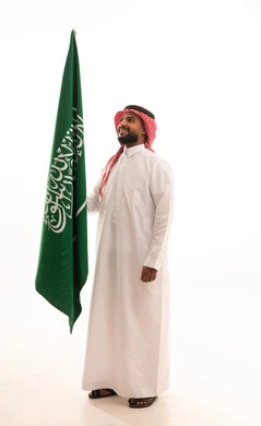 يوم العلم 11مارس، الاحتفال بذكرى اليوم الوطني السعودي 8 ربيع الأول, الفخر و الاعتزاز بالانتماء للمملكة, بورتريه لرجل عربي سعودي خليجي مبتسم يرتدي الشماغ والثوب التقليدي يحمل بيده علم السعودية , الجسم بالطول الكامل، خلفية بيضاء
