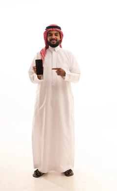 استعمال التقنيات الحديثة في التسويق الالكتروني، بورتريه لرجل عربي سعودي خليجي يرتدي الشماغ والثوب التقليدي يشير بيده الى شاشة اعلانية سوداء فارغة على الجوال، دور التقنية الحديثة في توفير راحة أكبر للأفراد، صورة الجسم بالطول الكامل، خلفية بيضاء