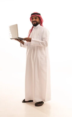 النظر الى الكاميرا بتعابير السعادة، اجراء مكالمة فيديو عبر الانترنت، إنجاز مهام العمل عن بعد, استخدام الأجهزة التقنية الحديثة والمتطورة, بورتريه لرجل عربي خليجي سعودي يرتدي الشماغ والثوب التقليدي يقوم باستعمال جهاز الحاسوب المحمول,  الجسم بالطول الكامل، خلفية بيضاء
