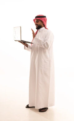 Raising the hand with expressions of greeting and peace, making a video call over the internet, completing work tasks remotely, using modern and advanced technological devices, a portrait of an Arab Gulf Saudi man wearing a ghutrah and traditional thobe using a laptop, full-body, white background.