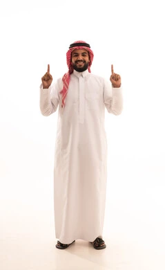 تحديد موقع شيء معين، حركات يدين واشارات تدل على هذا المكان (هنا)، بورتريه لرجل عربي خليجي سعودي مبتسم يرتدي الشماغ والثوب التقليدي يشير باصبعيه السبابة إلى الأعلى، الجسم بالطول الكامل، خلفيه بيضاء