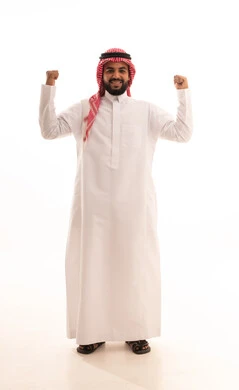 The concept of achievement and goal attainment, victory, winning, and success; a portrait of a smiling Arab Gulf Saudi man wearing a ghutrah and traditional thobe, looking at the camera, raising his fists up with gestures of joy and happiness, feeling of happiness and strength, full-body shot, white background.