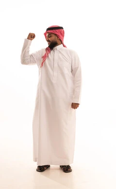 Physical fitness and health, showcasing physical strength, a portrait of a smiling Arab Gulf Saudi man wearing a ghutrah and traditional thobe, raising his hand with a clenched fist, the concept of muscular strength and muscle display, white background.