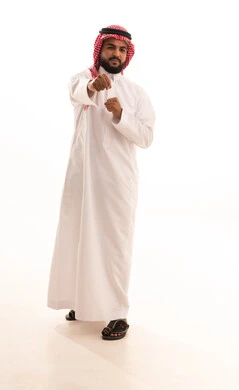 Mastering the arts of boxing, enhancing bone and joint health, practicing self-defense sports, increasing muscle endurance, a portrait of an Arab Gulf Saudi man wearing traditional attire and a ghutrah raising his fists in gestures of strength, full body length, white background.