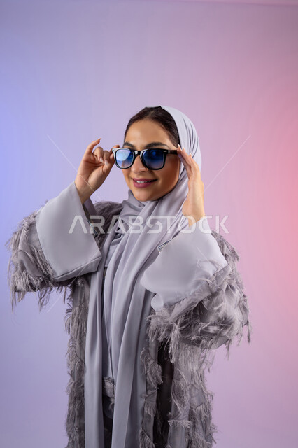 مفهوم الانوثة والأناقة والجمال, الاستمتاع بامتلاك اكسسوارات عصرية, سيدة اماراتية ترتدي عباءة ونظارة تنظر إلى الكاميرا بتعابير السرور, بورتريه لامرأة عربية خليجية سعودية محجبة تقوم بتجربة النظارات الشمسية الجديدة, خلفية ملونة