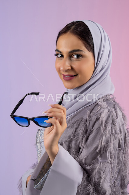 الاستمتاع بامتلاك اكسسوارات عصرية, الاهتمام بالاناقة والمظهر الخارجي، سيدة اماراتية ترتدي عباءة ونظارة, إيماءات الانوثة والجمال, بورترية مقرب لامرأة عربية خليجية سعودية محجبة تقوم بتجربة النظارات الشمسية الجديدة, النظر إلى الكاميرا بتعابير السرور، خلفية ملونة